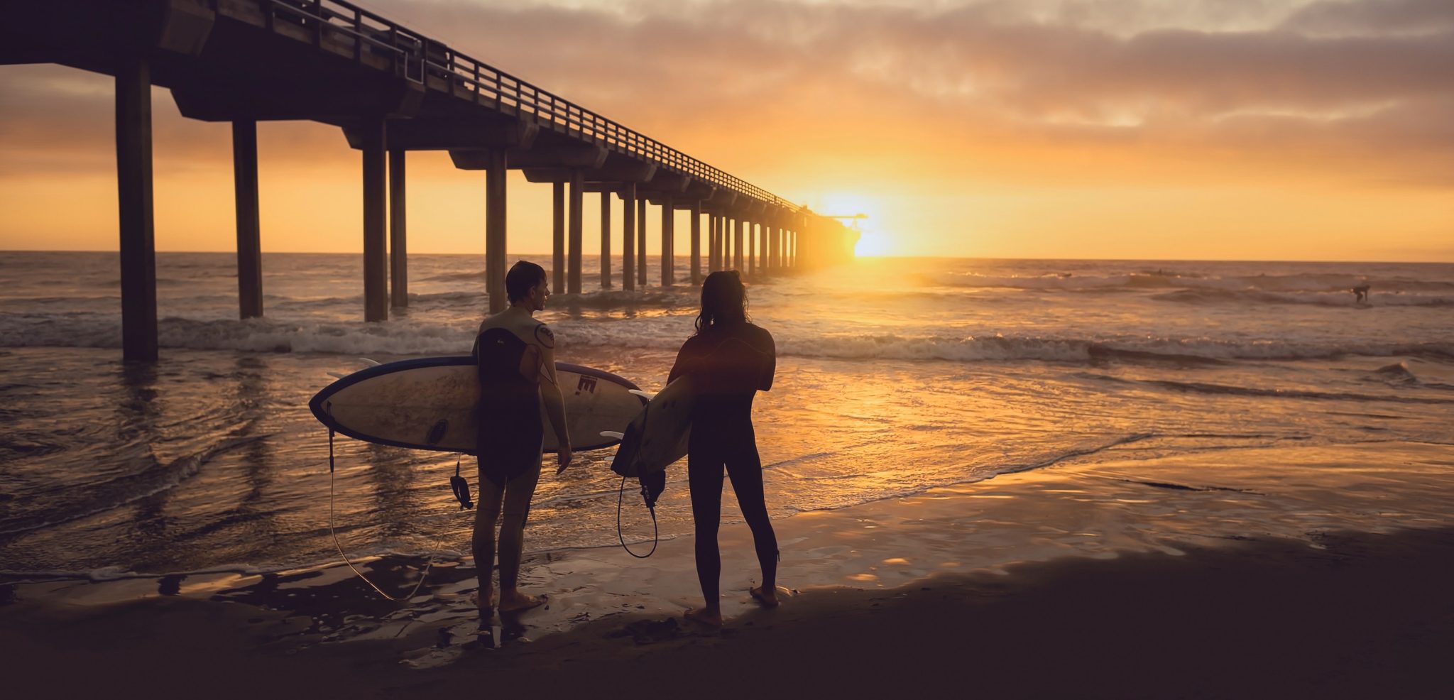 2 people, one holding a surfboard, looking out at the ocean at sunset